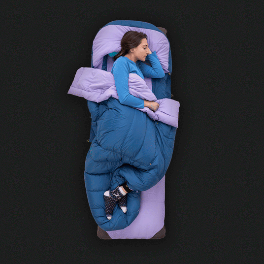 A person comfortably sleeping on their side in a Zenbivy bed setup. The bed includes a purple mattress sheet, blue down quilt, and matching hood, all laid out on a black background. The sleeper is wearing cozy socks and a blue long-sleeve shirt.
