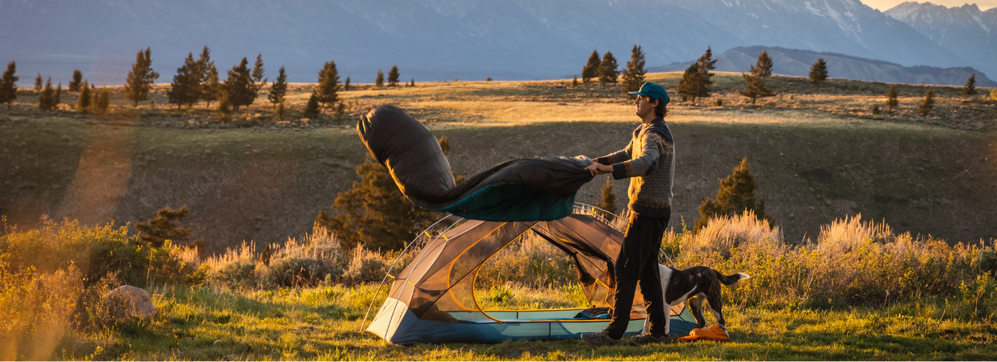 Person with a dog standing next to a tent in a field with mountains in the background. I zenbivy.eu